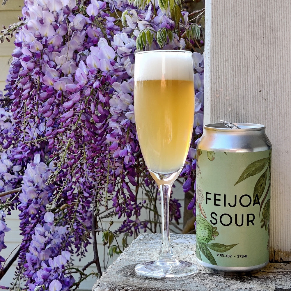 This is a photo of a green can of beer that has been made by Ocho and it is called Fiejoa Sour. The can of beer has been poured into a beer glass and the photo has a backdrop of some beautiful flowers