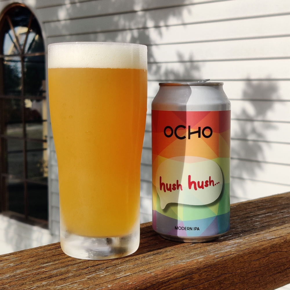 A photo of a can and a schooner glass of Hush Hush Session IPA beer. The glass is full of beer and you can see the brewery building in the back ground
